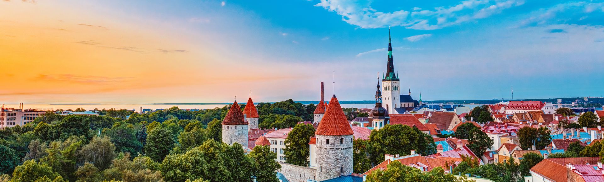 A picturesque view of Tallinn's skyline, featuring medieval towers with red roofs, a tall spire, lush greenery, and a blue sky at sunset. Tranquil and historic.