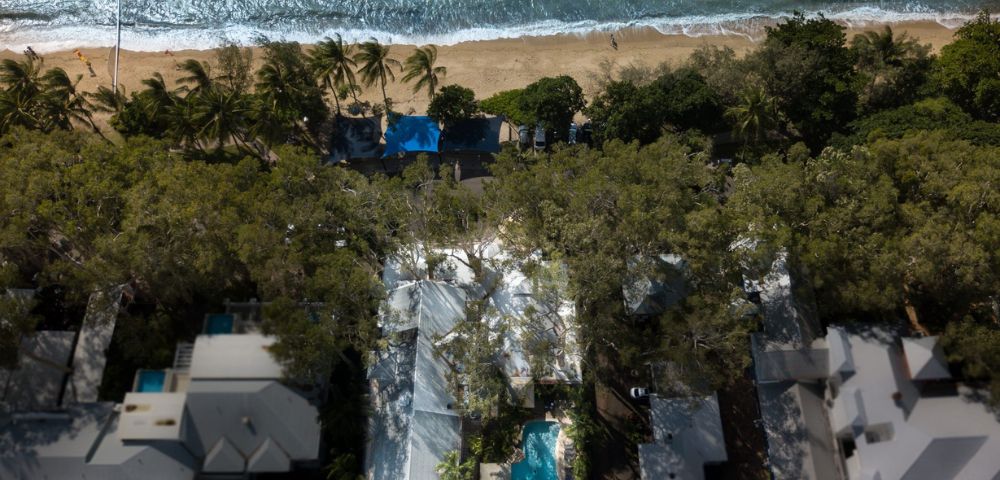 Aerial view of a coastal resort, featuring a lush green canopy, sandy beach with gentle waves, surrounded by elegant white buildings and a central pool.