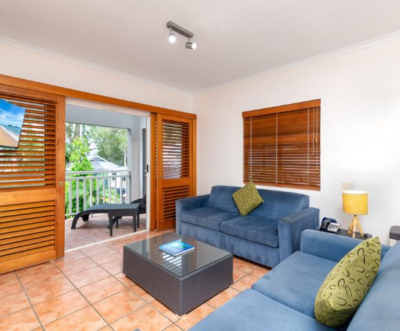 Cozy living room with blue couches, a glass coffee table, and a small lamp. Wooden doors open to a balcony with outdoor seating. Warm, inviting atmosphere.