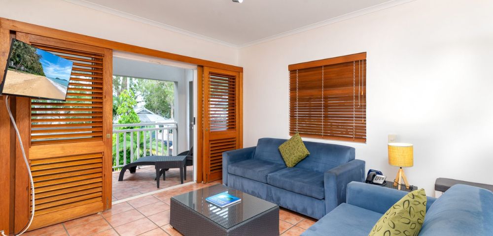 Cozy living room with blue couches, a glass coffee table, and a small lamp. Wooden doors open to a balcony with outdoor seating. Warm, inviting atmosphere.