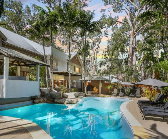 Idyllic tropical resort pool scene with clear blue water, surrounded by lush palm trees and lounge chairs. The sky has a warm, sunset glow.