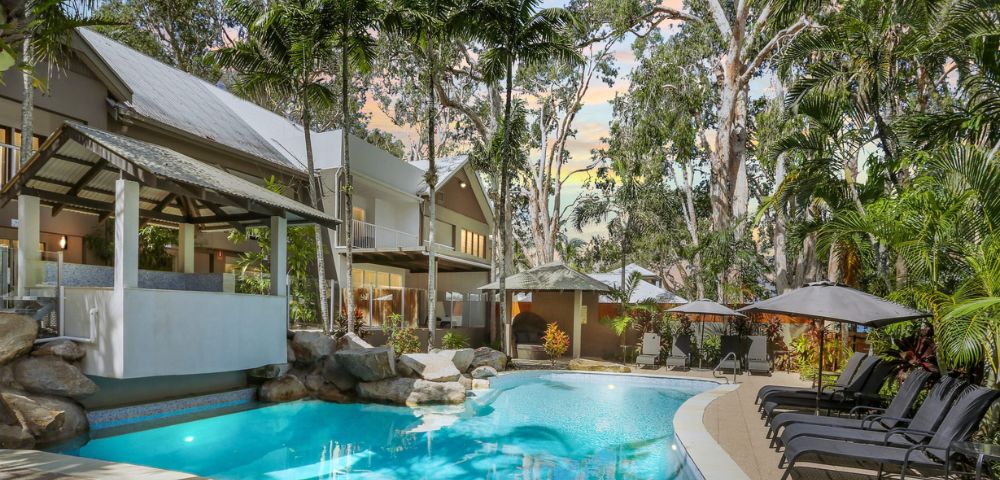 Idyllic tropical resort pool scene with clear blue water, surrounded by lush palm trees and lounge chairs. The sky has a warm, sunset glow.
