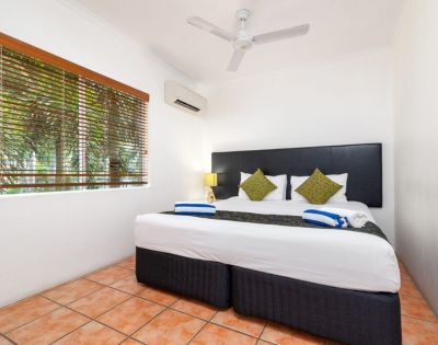 A cozy hotel room with a large bed, two green pillows, and a modern headboard. There are blue-striped towels, a bedside lamp, and a window with blinds.