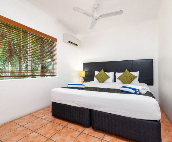 A cozy hotel room with a large bed, two green pillows, and a modern headboard. There are blue-striped towels, a bedside lamp, and a window with blinds.
