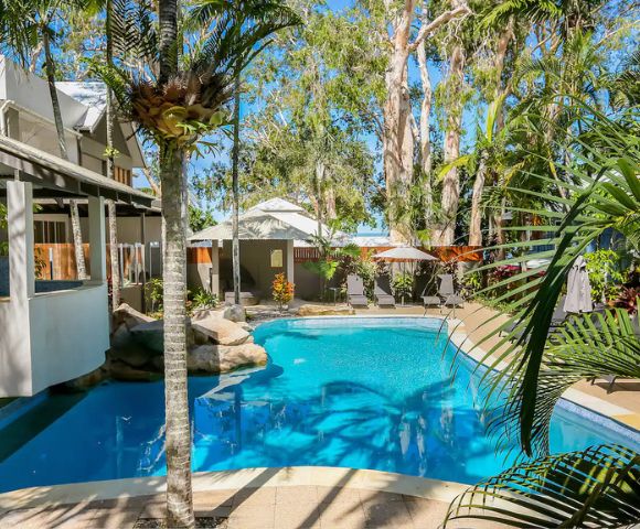 A tranquil outdoor pool surrounded by tall trees and lush plants, adjacent to a building with white verandas, under a clear blue sky. Relaxing and inviting.