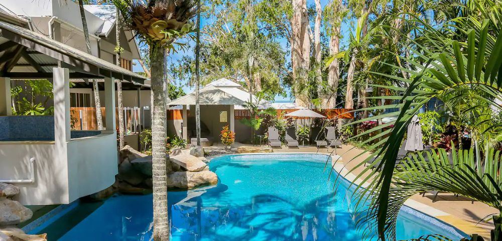 A tranquil outdoor pool surrounded by tall trees and lush plants, adjacent to a building with white verandas, under a clear blue sky. Relaxing and inviting.