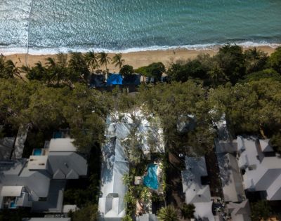Aerial view of a coastal resort, featuring a lush green canopy, sandy beach with gentle waves, surrounded by elegant white buildings and a central pool.