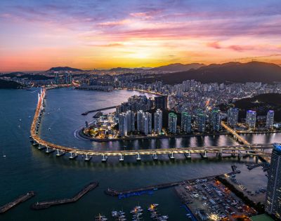 Aerial view of a coastal city at sunset with a brightly lit bridge spanning a wide river. Skyscrapers line the water, and mountains are silhouetted against a vibrant sky.