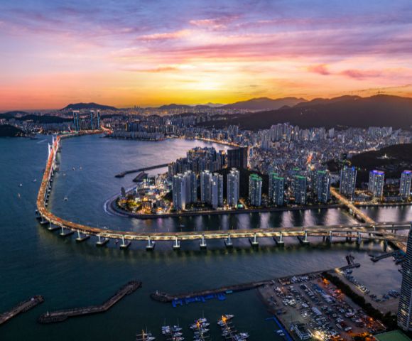 Aerial view of a coastal city at sunset with a brightly lit bridge spanning a wide river. Skyscrapers line the water, and mountains are silhouetted against a vibrant sky.
