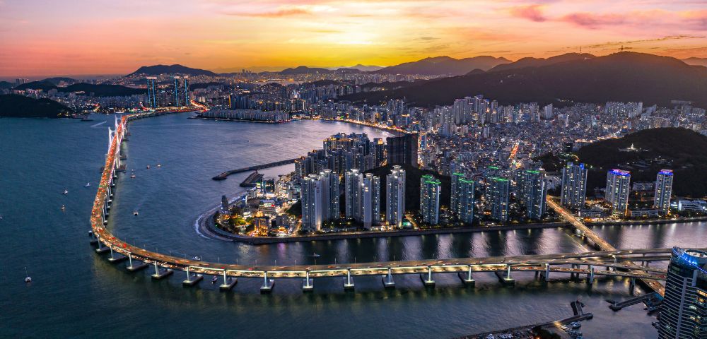 Aerial view of a coastal city at sunset with a brightly lit bridge spanning a wide river. Skyscrapers line the water, and mountains are silhouetted against a vibrant sky.