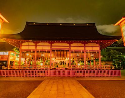 Traditional Japanese temple illuminated at night with warm lights. The structure has a curved roof and wooden details, surrounded by a pink fence, creating a serene and mystical atmosphere.