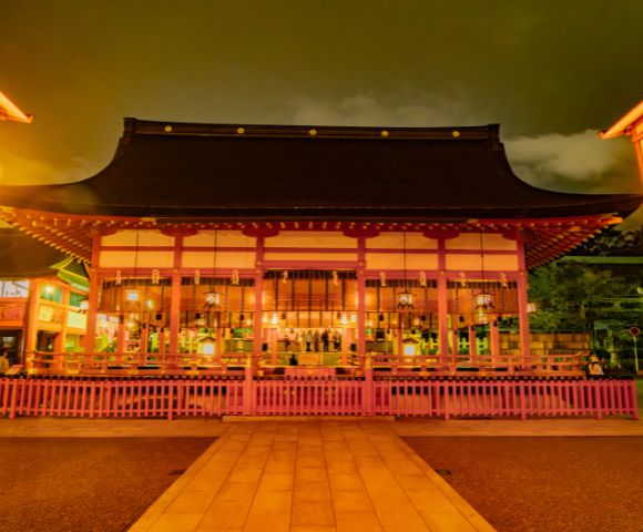 Traditional Japanese temple illuminated at night with warm lights. The structure has a curved roof and wooden details, surrounded by a pink fence, creating a serene and mystical atmosphere.