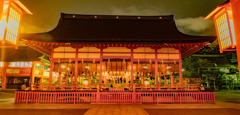 Traditional Japanese temple illuminated at night with warm lights. The structure has a curved roof and wooden details, surrounded by a pink fence, creating a serene and mystical atmosphere.