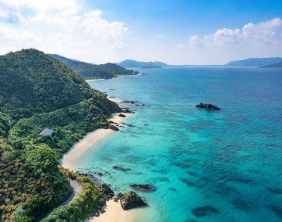 Aerial view of a serene coastline with lush green hills, a sandy beach, and turquoise ocean. The sky is clear and blue, evoking a sense of peace and tranquility.
