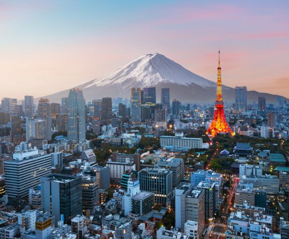 Aerial view of Tokyo at sunset with Mount Fuji in the background. The Tokyo Tower glows with warm lights, surrounded by urban skyscrapers under a pastel sky.