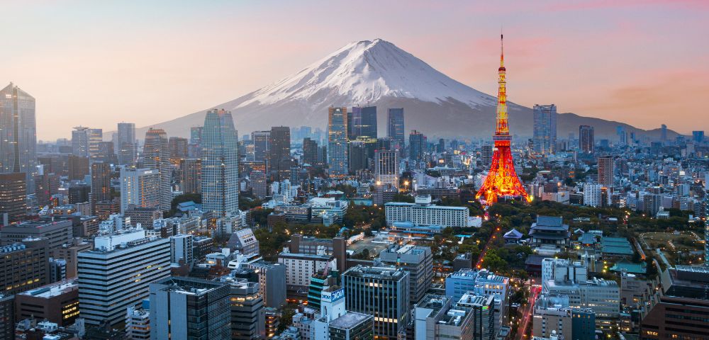 Aerial view of Tokyo at sunset with Mount Fuji in the background. The Tokyo Tower glows with warm lights, surrounded by urban skyscrapers under a pastel sky.