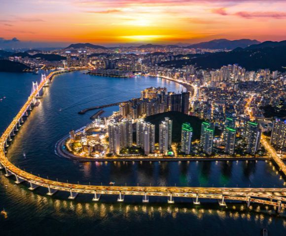 Aerial view of a vibrant coastal city at sunset, featuring illuminated skyscrapers, a bridge over water, and glowing skies, conveying a lively urban scene.