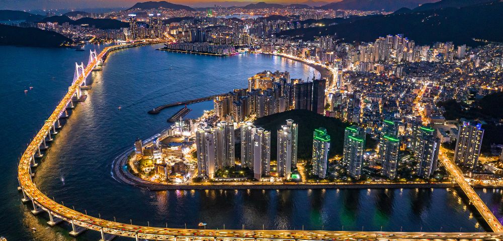 Aerial view of a vibrant coastal city at sunset, featuring illuminated skyscrapers, a bridge over water, and glowing skies, conveying a lively urban scene.
