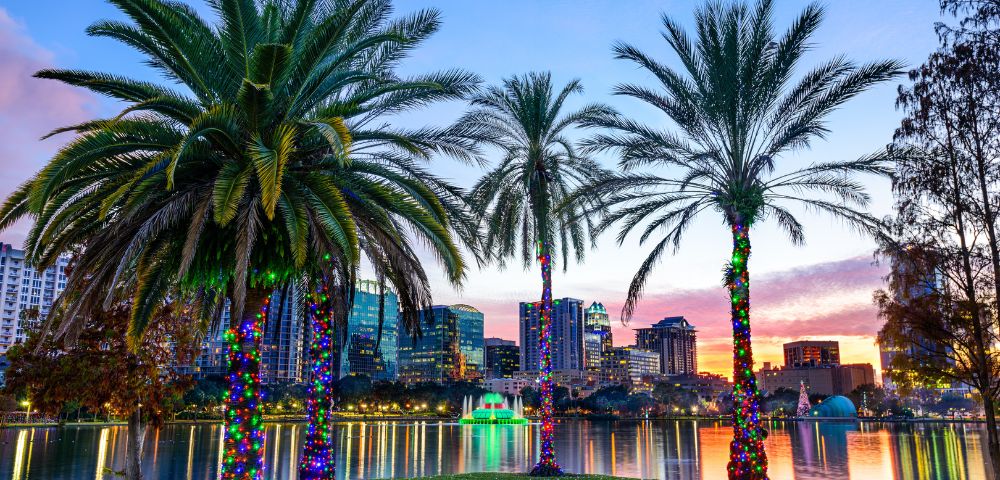 Twilight view of a cityscape with palm trees adorned in colorful lights by a lake. Reflections shimmer on the water, and the sky is a mix of oranges and purples.