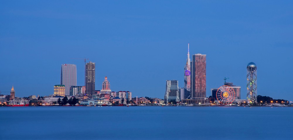 City skyline at dusk featuring modern high-rises and a ferris wheel, reflecting on calm water. The sky is a deep blue, creating a serene atmosphere.