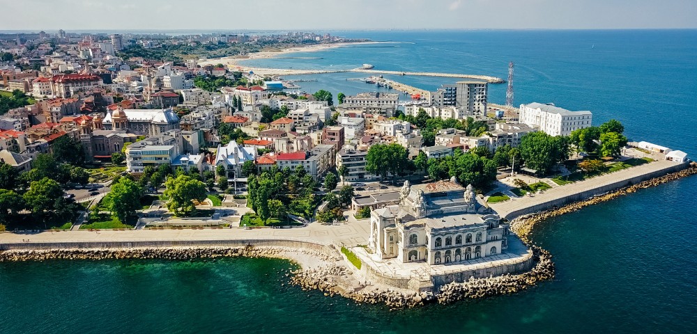 Aerial view of Constanța, Romania, showcasing the historical casino on the seafront. The vibrant cityscape extends to sandy beaches, under a clear blue sky.