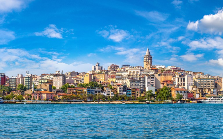 Scenic view of Istanbul's skyline with the historic Galata Tower rising above colorful buildings. A calm blue sea and a clear sky enhance the lively atmosphere.