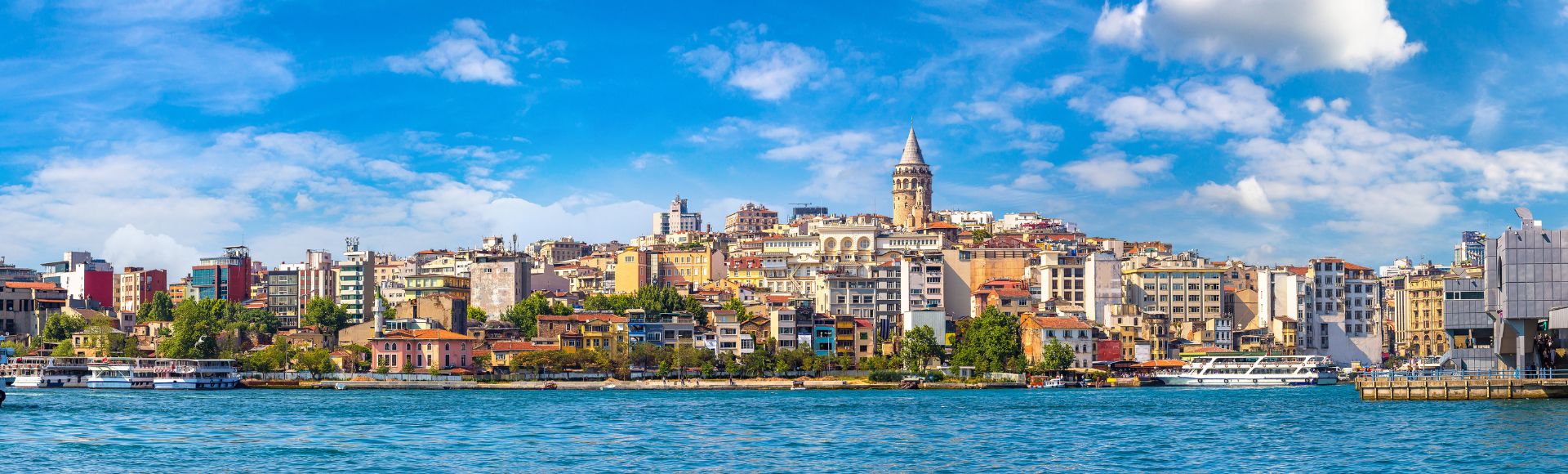 Scenic view of Istanbul's skyline with the historic Galata Tower rising above colorful buildings. A calm blue sea and a clear sky enhance the lively atmosphere.