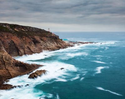 Rocky coastline with jagged cliffs meeting the ocean, waves crashing against them. Overcast sky adds a dramatic, serene mood to the scene.