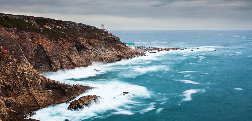 Rocky coastline with jagged cliffs meeting the ocean, waves crashing against them. Overcast sky adds a dramatic, serene mood to the scene.