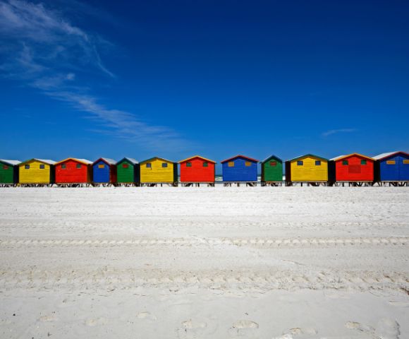 Colorful beach huts in red, yellow, green, and blue line a bright, sandy beach under a clear blue sky, evoking a cheerful, summer atmosphere.