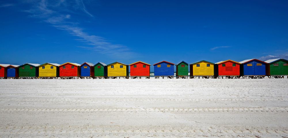 Colorful beach huts in red, yellow, green, and blue line a bright, sandy beach under a clear blue sky, evoking a cheerful, summer atmosphere.