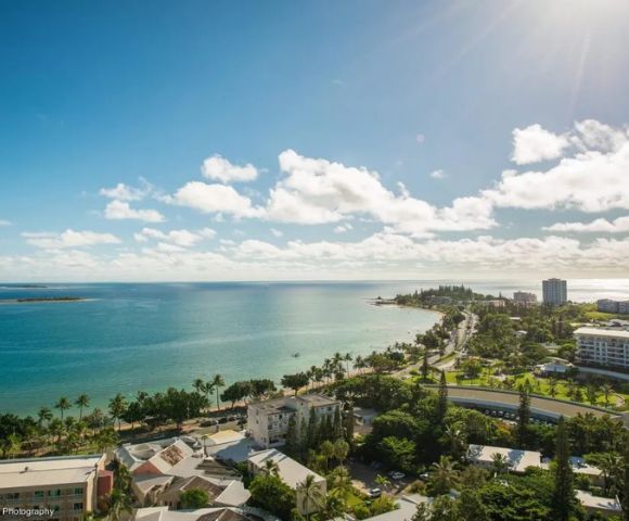 Aerial view of a coastal city with blue ocean under a bright, sunny sky. Lush greenery lines the shore, buildings scattered throughout, conveying a serene, vibrant atmosphere.