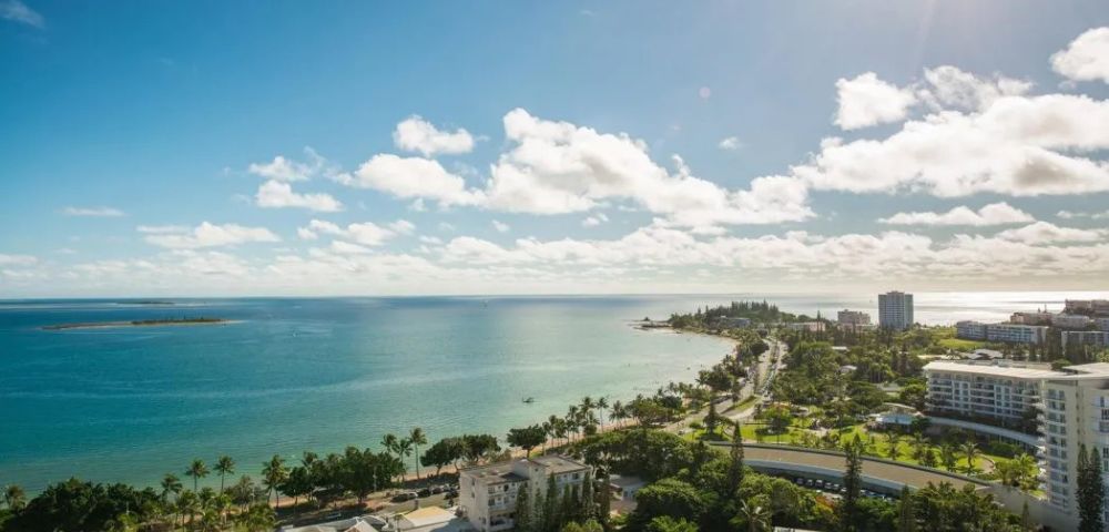 Aerial view of a coastal city with blue ocean under a bright, sunny sky. Lush greenery lines the shore, buildings scattered throughout, conveying a serene, vibrant atmosphere.