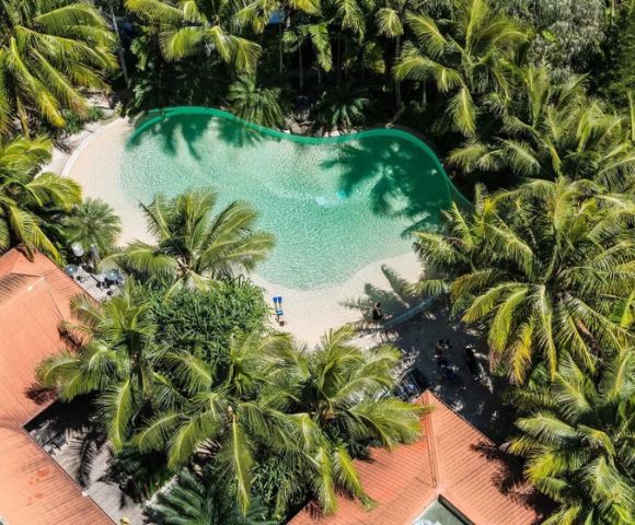 Aerial view of a lagoon-shaped swimming pool surrounded by lush palm trees and red-roofed buildings, conveying a serene tropical oasis vibe.