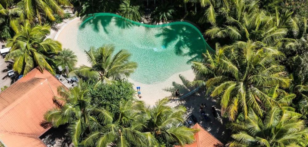 Aerial view of a lagoon-shaped swimming pool surrounded by lush palm trees and red-roofed buildings, conveying a serene tropical oasis vibe.