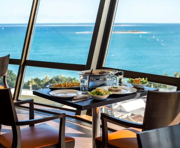 Dining table by large windows with a view of the ocean. Plates and bowls with food, two chairs, and bright blue sea create a serene and inviting atmosphere.