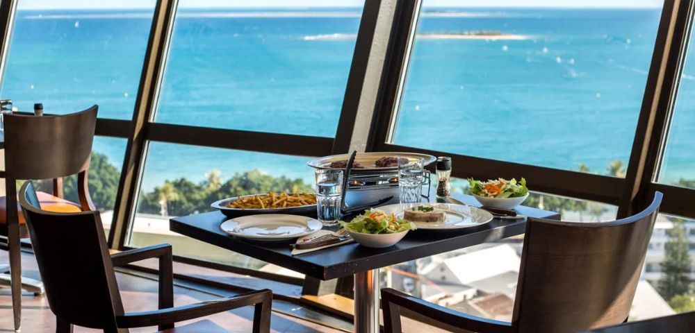 Dining table by large windows with a view of the ocean. Plates and bowls with food, two chairs, and bright blue sea create a serene and inviting atmosphere.