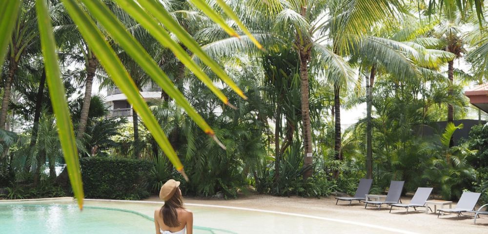 Woman in a white bikini stands at the edge of a serene pool surrounded by lush tropical trees. Sun loungers line the background, conveying a peaceful, vacation vibe.