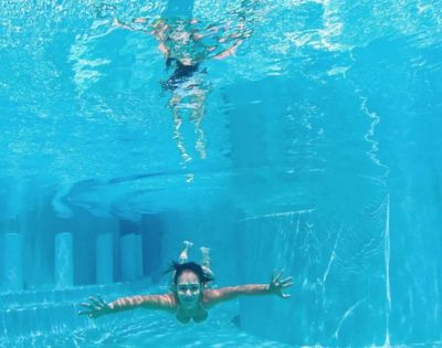 A person swims underwater in a clear pool, facing forward with arms outstretched. Sunlight creates wavy patterns on the surface, conveying a serene, playful mood.