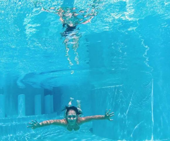 A person swims underwater in a clear pool, facing forward with arms outstretched. Sunlight creates wavy patterns on the surface, conveying a serene, playful mood.