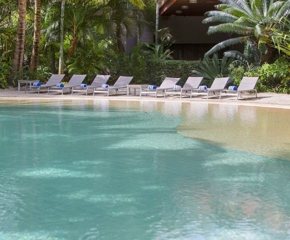 Serene poolside scene with turquoise water, surrounded by lush greenery. Multiple empty lounge chairs line one side, evoking a peaceful atmosphere.