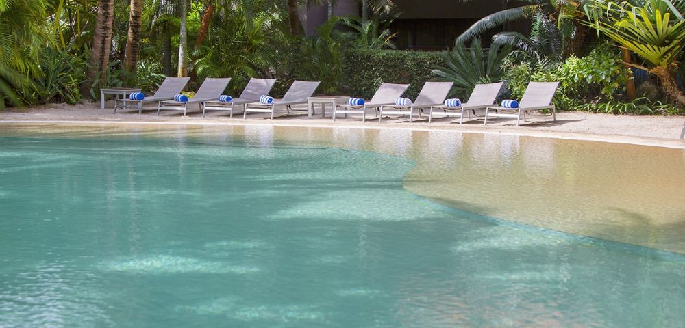 Serene poolside scene with turquoise water, surrounded by lush greenery. Multiple empty lounge chairs line one side, evoking a peaceful atmosphere.