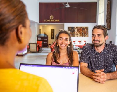A couple smiles warmly at a concierge desk, with a laptop open in the foreground. The setting is welcoming, with modern decor and warm lighting.
