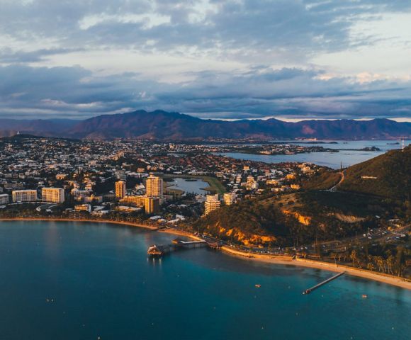Aerial view of a coastal city flanked by a serene bay and rolling hills. Buildings line the shore under a sky streaked with clouds, evoking a tranquil evening.