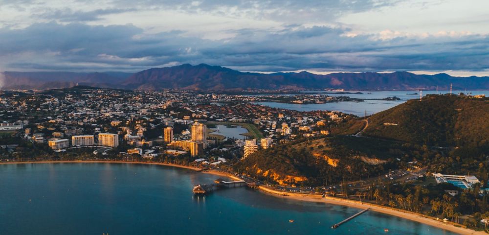 Aerial view of a coastal city flanked by a serene bay and rolling hills. Buildings line the shore under a sky streaked with clouds, evoking a tranquil evening.