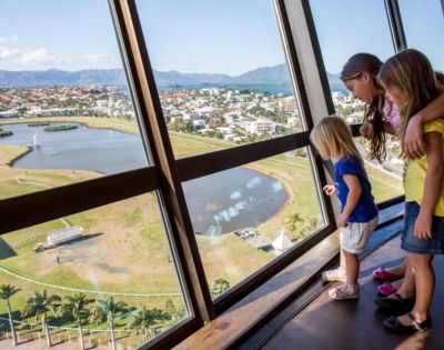 Three children look out a tilted window with a scenic view of a city, lake, and mountains. The atmosphere is curious and peaceful.