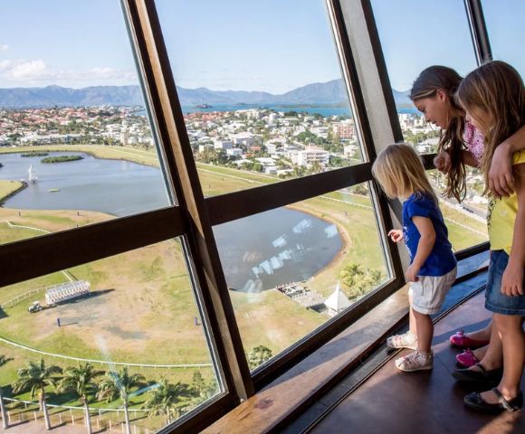 Three children look out a tilted window with a scenic view of a city, lake, and mountains. The atmosphere is curious and peaceful.