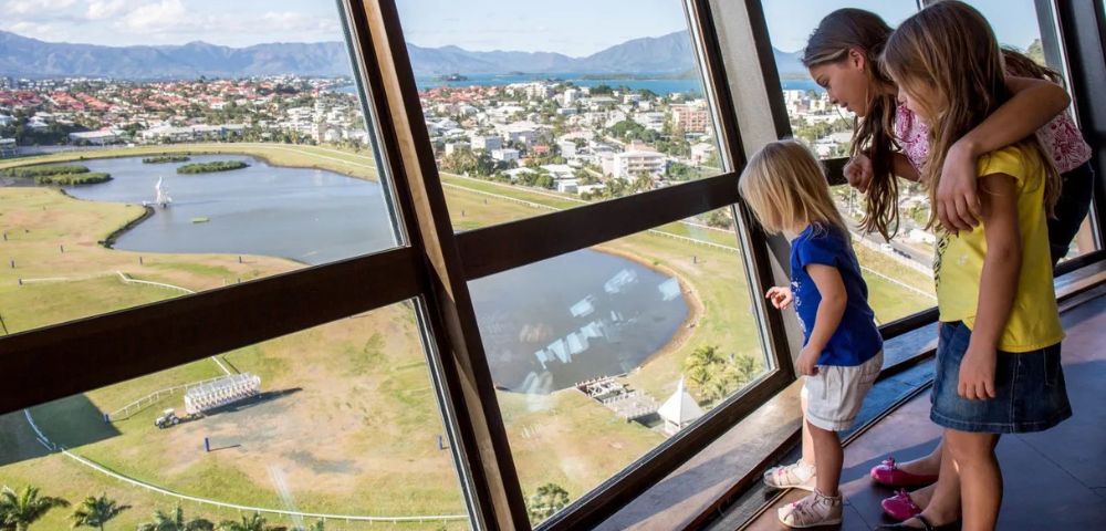 Three children look out a tilted window with a scenic view of a city, lake, and mountains. The atmosphere is curious and peaceful.