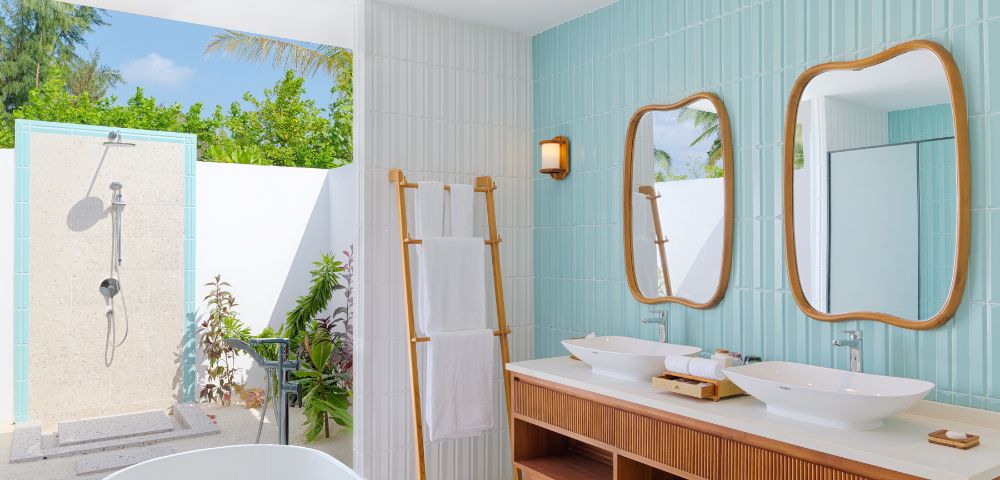 Modern outdoor bathroom with a tropical feel. Features twin wood-framed mirrors, double sinks, a freestanding tub, and an open-air shower. Bright and serene.