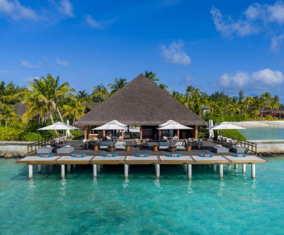 A tropical resort with thatched-roof building on stilts over turquoise water. Surrounding the building are palm trees and clear skies, evoking relaxation.